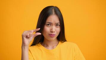 A millennial woman with long hair playfully makes a gesture with her hand, showing a small size. She stands in front of a vibrant orange backdrop, adding energy to the moment. video