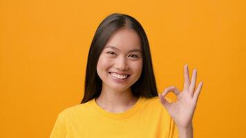 A joyful Asian woman in a yellow shirt smiles while making an OK gesture with her right hand. The vibrant yellow background highlights her cheerful expression and youthful energy. video
