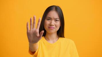 A millennial woman with long hair stands in front of a vibrant yellow backdrop, raising her hand in a stop gesture. She conveys a message that suggests pause or rejection. video