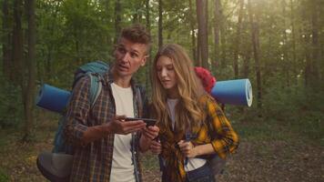 A couple stands together in a vibrant forest, engaged with a smartphone as they take a break from hiking. Their backpacks rest behind them, and the sun filters through the trees. video