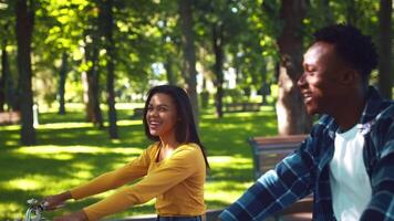 A young African American couple is cycling together through a lush green park on a sunny day. Their joyful expressions highlight their connection and enjoyment of the outdoors. video