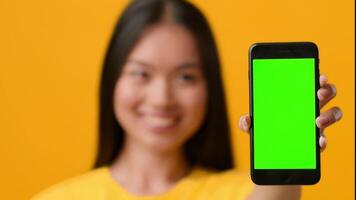 A young Asian woman smiles while showcasing a smartphone with a blank green screen. She stands against a vibrant yellow backdrop, reflecting a modern lifestyle and technology focus. video