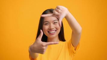 A young Asian woman joyfully frames her face with her hands, showcasing creativity against a vibrant yellow backdrop. Her expression radiates confidence and enthusiasm for self-expression. video
