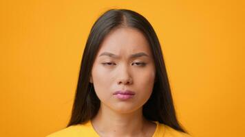 A young Asian woman displays a serious expression against a vibrant yellow backdrop. She is dressed casually, embodying a modern lifestyle while looking directly at the viewer. video