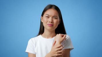 A young Asian woman stands against a blue background, showing a pained expression while holding her wrist. She appears to be experiencing discomfort in a relaxed atmosphere. video