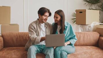 A young couple sits together on a cozy sofa, reviewing real estate listings on a laptop amidst moving boxes in their new apartment. They appear happy and excited about their relocation. video