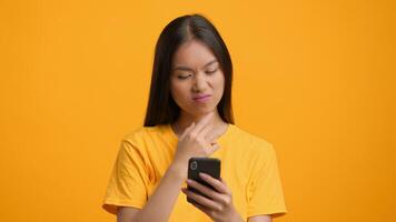 A millennial woman of Asian descent focuses on her smartphone, dressed in a bright yellow shirt. The vivid background enhances her modern lifestyle while she engages with technology. video