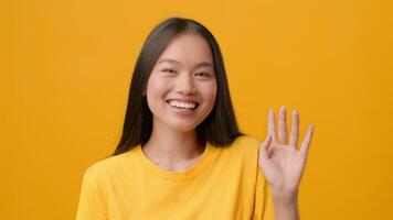 A young Asian woman with long hair smiles brightly and waves her hand. She wears a cheerful yellow shirt, creating a vibrant atmosphere that captures a moment of joy and positivity. video
