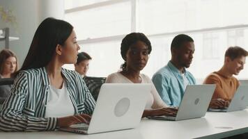A group of diverse students is focused on their laptops during a study session at a university. They collaborate in a bright, modern classroom filled with natural light. video