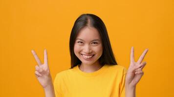 A joyful woman with long dark hair poses against a vibrant yellow backdrop, happily showing a peace sign with both hands. Her bright yellow shirt complements the lively atmosphere. video