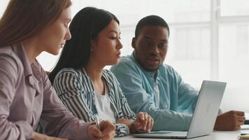 Three diverse students engage in a focused discussion while studying together at a university. They work on a laptop, sharing ideas and taking notes in a modern, well-lit environment. video