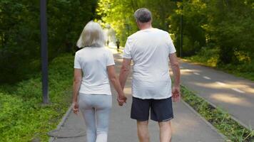 A senior couple in their 60s walks together in a lush green park, holding hands as they enjoy their exercise routine. The atmosphere is bright and inviting, showcasing their active lifestyle. video