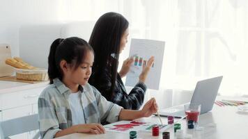 In a bright kitchen, a mother and daughter share a joyful moment creating art. The daughter paints while the mother working online, surrounded by colorful materials and a laptop. video