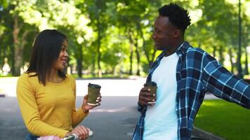 A young African American couple smiles and engages in conversation while holding coffee cups. They are surrounded by greenery in a lively park, enjoying each other's company on a sunny day. video