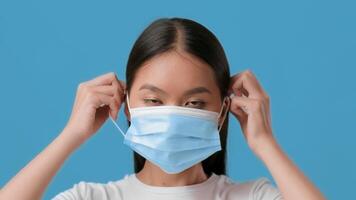 A millennial Asian woman is adjusting a light blue mask on her face against a solid blue background. She demonstrates caution and health awareness in her daily routine. video