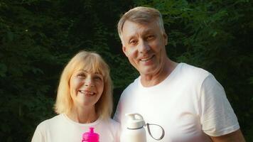 In a lush park setting, a senior couple in their 60s smiles while holding water bottles. They promote an active lifestyle through jogging and outdoor exercise together, embracing their health. video
