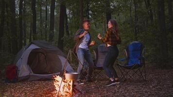 A couple engages in playful dancing by a campfire at their forest campsite. Surrounded by trees, they enjoy their time outdoors, showcasing a vibrant and active lifestyle beneath the stars. video