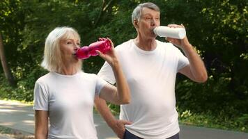 A senior couple is taking a break from their jogging session in a scenic park. They are sipping water from colorful bottles while enjoying the fresh air and greenery around them. video