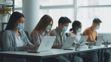 diverso grupo do alunos ativamente estudando com laptops dentro uma brilhante, moderno sala de aula. elas vestem máscaras, destacando atenção para segurança enquanto perseguindo Educação dentro uma universidade contexto. video