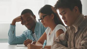 Three college students participate in a dedicated study session, taking notes and concentrating on their work in a bright, modern classroom. Their expressions reflect determination and focus. video
