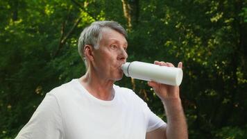 A senior man jogs in a lush green park, maintaining an active lifestyle. He stop briefly to hydrate, enjoying the fresh air and beautiful scenery surrounding video