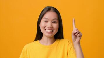A cheerful Asian woman is isolated against a vibrant yellow backdrop, smiling and raising her finger as if to share an idea or suggestion. Her casual attire complements the lively atmosphere. video