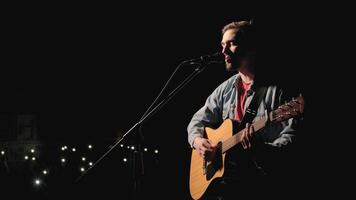 A musician is playing guitar on a large stage with audience members holding flashlights in the background. The auditorium is filled with beautiful flashing lights. 19 video