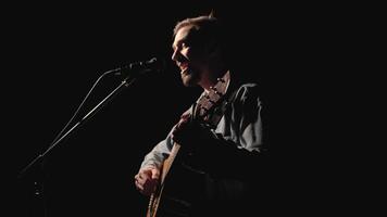 A musician is playing guitar on a large stage with audience members holding flashlights in the background. The auditorium is filled with beautiful flashing lights. 26 video