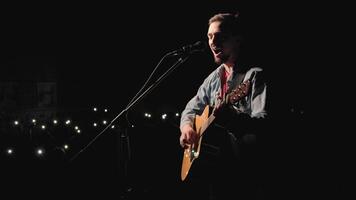 A musician is playing guitar on a large stage with audience members holding flashlights in the background. The auditorium is filled with beautiful flashing lights. 15 video