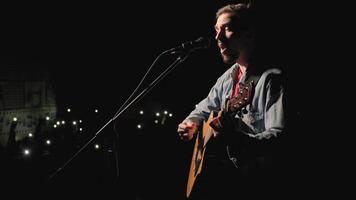 A musician is playing guitar on a large stage with audience members holding flashlights in the background. The auditorium is filled with beautiful flashing lights. 13 video