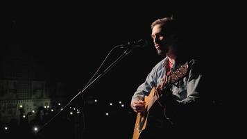 A musician is playing guitar on a large stage with audience members holding flashlights in the background. The auditorium is filled with beautiful flashing lights. 18 video