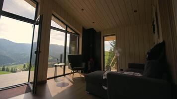 Cozy modern cabin with panoramic mountain view. Interior of a stylish wooden cabin with large glass doors open to a deck, offering stunning views of green video