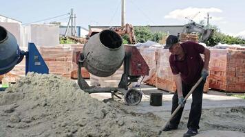 Construction worker mixing sand near cement mixer. Older man at construction site using a shovel to mix sand in front of a concrete mixer, with bricks and video