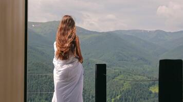 Woman wrapped in blanket enjoying mountain view. Young woman wrapped in a white blanket stands on a terrace, peacefully admiring the lush green mountain video