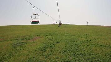 Empty ski lift chairs over grassy hillside. Red and green ski lift chairs ascend a green, open hillside under a hazy sky during the off-season in a mountain video