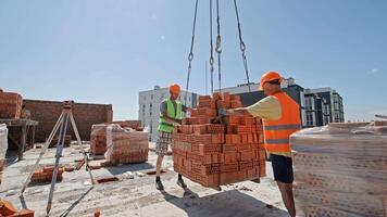 Construction workers guiding suspended brick pallet. Two construction workers in safety gear guide a crane-lifted pallet of perforated bricks at a building site video