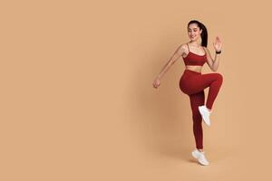 Warmup. Full body length shot of smiling young fitness model in sportswear standing on one leg and touching knee with her elbow, doing high knees exercise isolated over pastel studio background photo