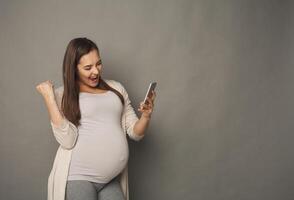 Surprised pregnant woman using smartphone copy space. Excited expectant lady with open mouth looking at mobile phone, gray studio background. Success, modern technologies, pregnancy concept photo