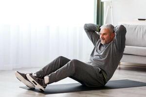 Athletic bearded senior man in grey sportswear doing exercises for abs at home, sitting on fitness mat and crunching, copy space. Healthy lifestyle, sport on self-isolation for older people photo