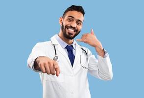 Cheerful Arab Doctor Male Gesturing Call Me Holding Hand Like Cellphone Near Ear And Pointing Finger To Camera Posing On Blue Studio Background, Smiling To Camera. Medical Consultation By Phone photo
