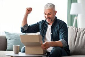 Happy senior man customer checking mail box, raising fists up and screaming, sitting on sofa at home, copy space. Emotional elderly man opening delivery, checking order. Delivery concept photo