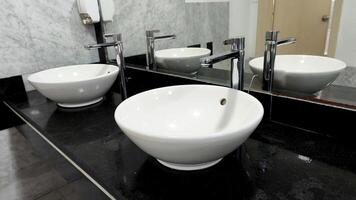 Sleek white vessel sinks with tall chrome faucets on a polished black counter, reflected in a large mirror inside a modern, minimalistic restroom interior. photo