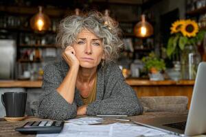 Middle aged woman sits at kitchen with worried look, surrounded by documents, calculator, and laptop photo