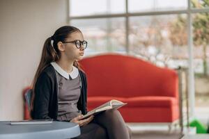 Young girl in uniform and glasses reading book at round table in bright interior with red sofa background reflecting calm learning moment modern aesthetic intelligence and soft visual storytelling photo