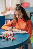 Creative girl painting with concentration at colorful art table surrounded by paints and brushes wearing orange sweater showing mindful childhood creativity and emotional development through art photo
