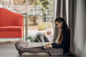 Teen girl in glasses working on laptop while sitting on floor near window with natural light soft curtains and minimalist background showing calm indoor lifestyle focused digital routine and remote photo
