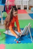 Girl painting at wooden easel on colorful foam mat using brush and jars of paint in creative classroom showing independence imagination and peaceful concentration during early childhood artistic photo