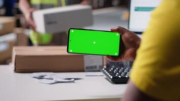 Staff reviews green screen pc in the storage room for data entry, inventory tracking mockups and visuals or barcode printer interfaces. Logistics worker packages cargo in the foreground. Camera B. video