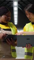 Vertical Team of diverse workers handling tracking software for parcel status on tablet, following the cargo shipment from the industrial warehouse with racks. Staff in hi vis vests. Camera B. video