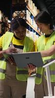 Vertical Team of diverse workers handling tracking software for parcel status on tablet, following the cargo shipment from the industrial warehouse with racks. Staff in hi vis vests. Camera A. video